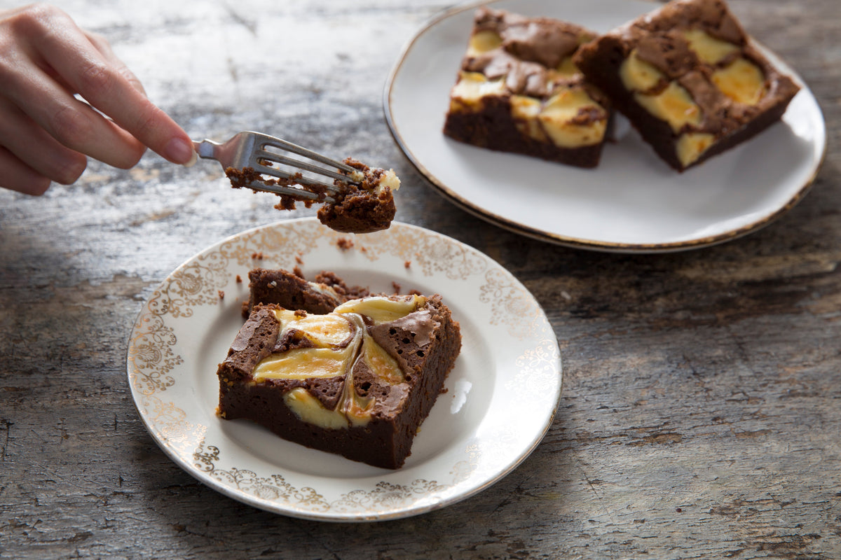 Vanilla cheesecake brownies with golden vanilla swirls on white plates, with a fork lifting a bite.
