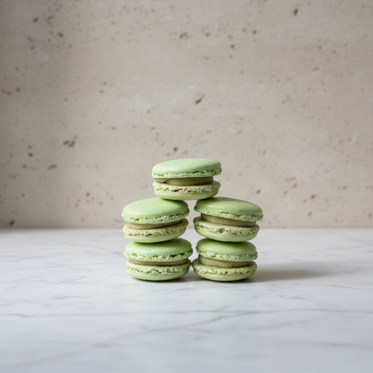 Light green pistachio macarons stacked in two columns on a rustic wooden surface, with a bamboo screen background.
