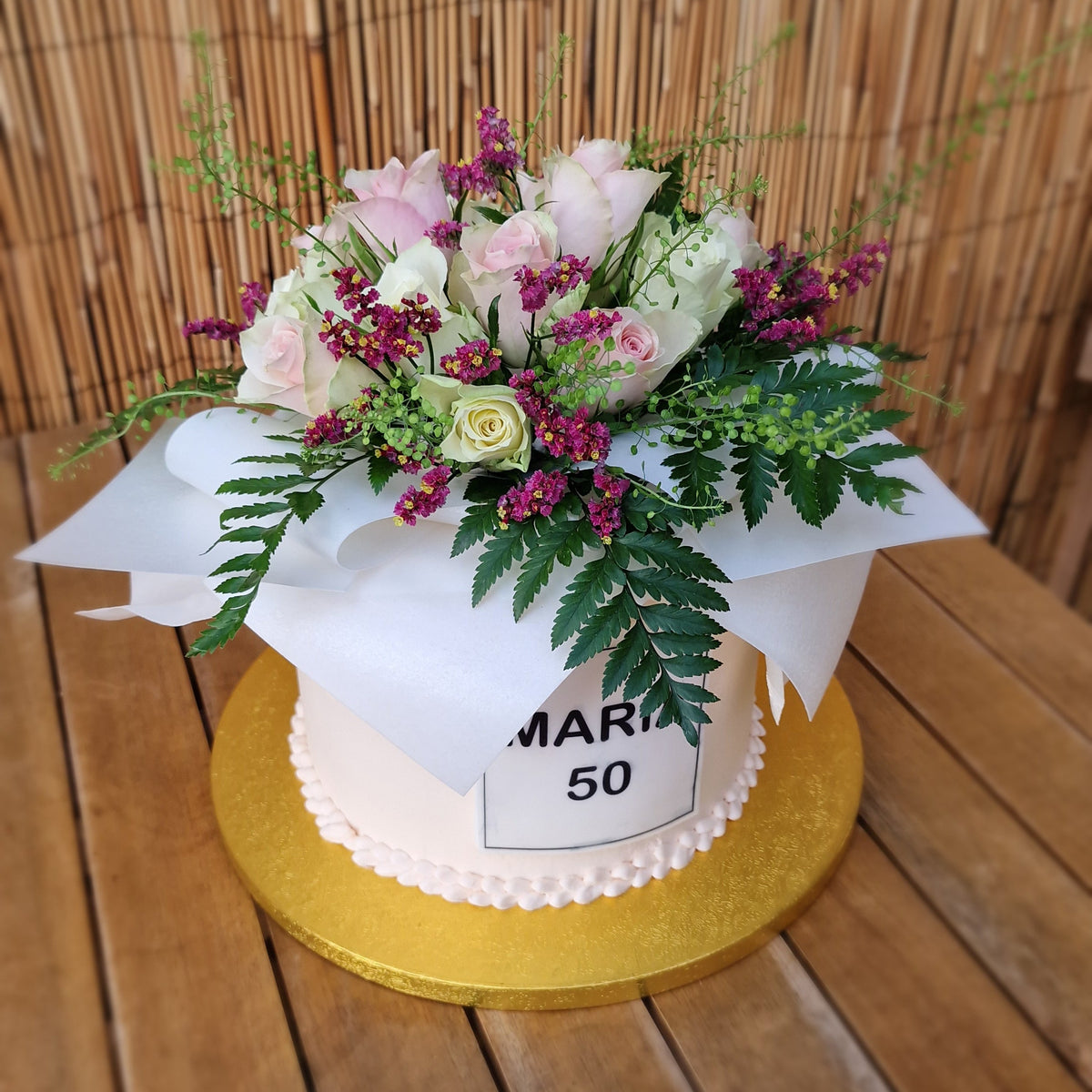 A white tiered cake on a gold round base, decorated with a floral arrangement of pink, white, and burgundy flowers with green foliage, wrapped in white tissue paper. The cake sits on a wooden table with a bamboo screen background.