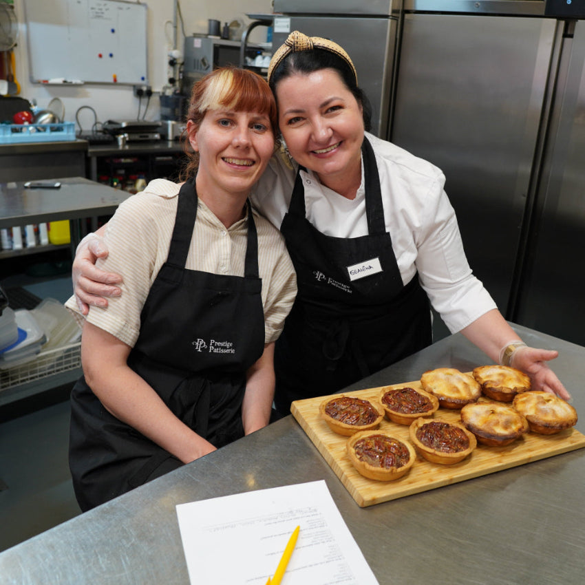Two women in black aprons standing in a commercial kitchen next to a wooden board holding several baked tartlets or pastries.