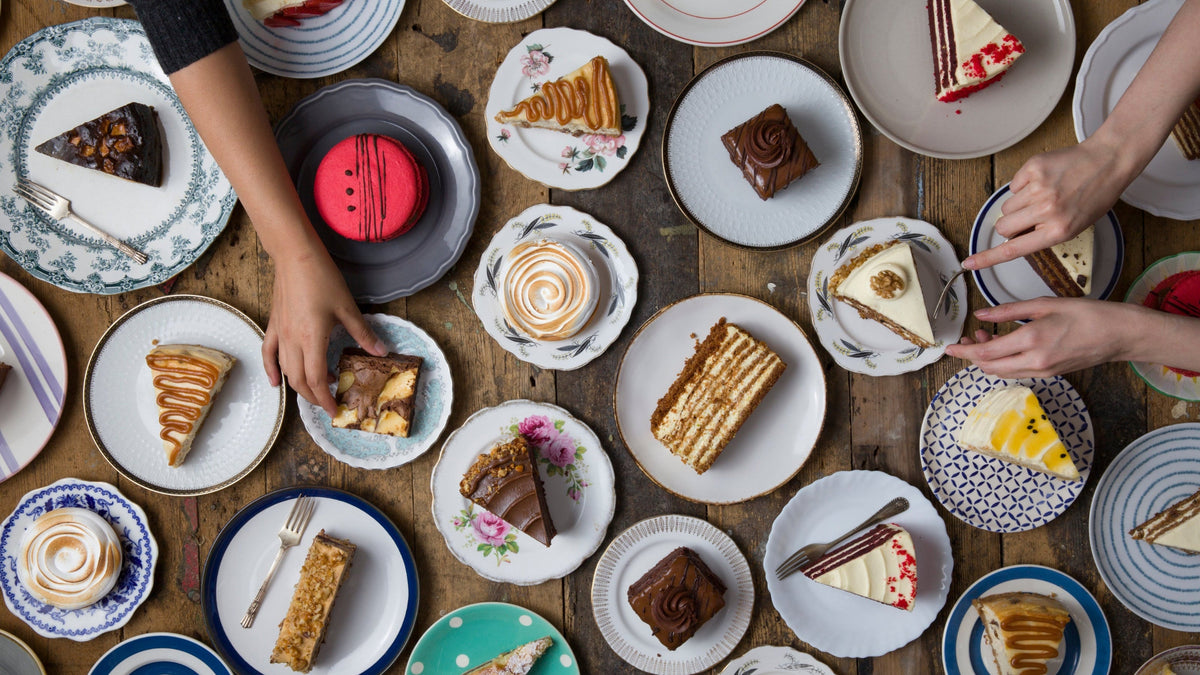 Tabletop scene with various desserts on plates, hands reaching for treats.
