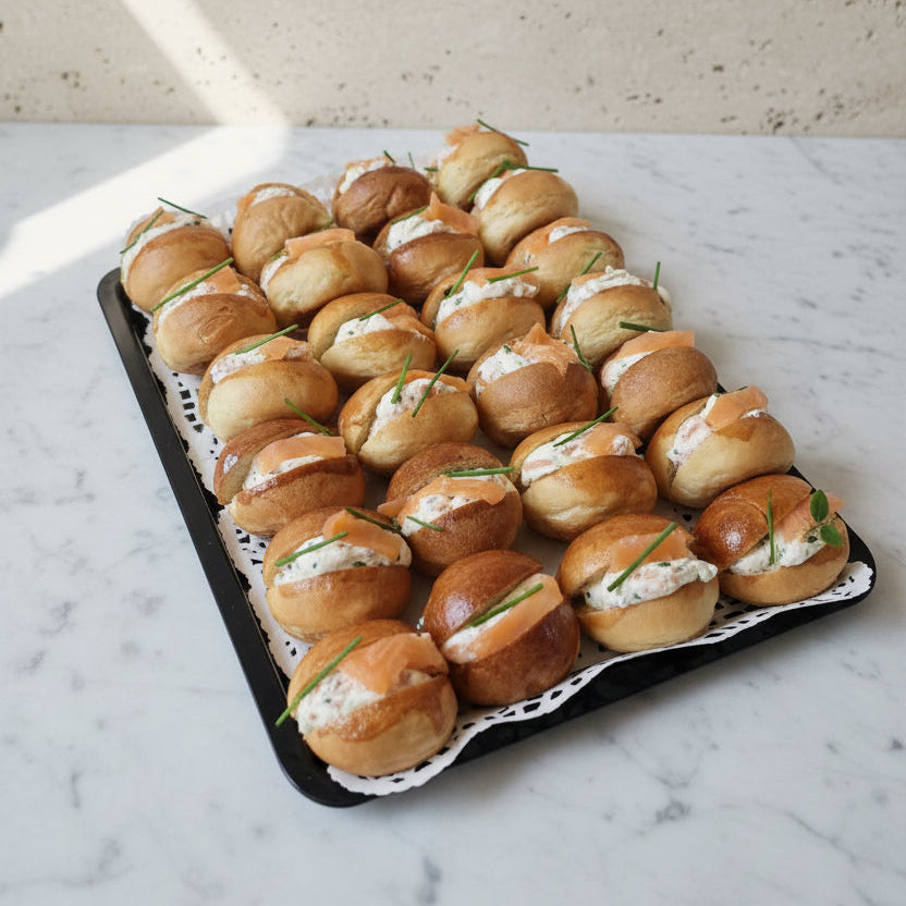 A tray of mini brioches filled with chive cream cheese ans smoked salmon, arranged on a white lace doily over a gray surface.