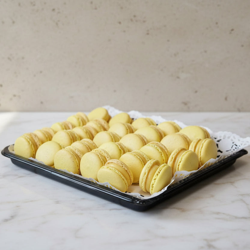 A black tray lined with a white lace doily, filled with rows of yellow passion fruit  macarons, set on a stainless steel countertop.